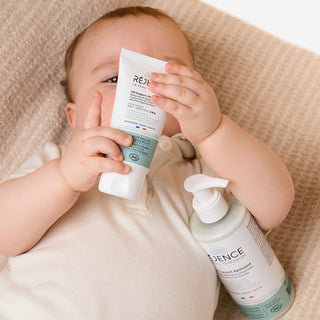 Bébé allongé tenant la Cold Cream SOS Réjence avec le soin lavant apaisant posé à ses côtés, illustrant la routine SOS eczéma bébé.