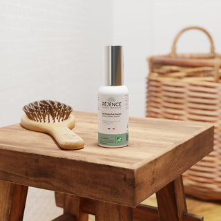 Flacon de l’Eau Florale Cuir Chevelu Réjence posé sur un meuble en bois à côté d’une brosse à cheveux, dans une salle de bain lumineuse aux tons naturels.