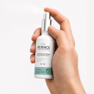 Gros plan d’une main tenant le flacon de l’Eau Florale Cuir Chevelu Réjence et appuyant sur le spray, avec une fine brume visible sur fond blanc.