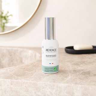 Flacon d’Eau Florale Purifiante Réjence posé sur un plan de toilette en pierre claire devant un miroir, à côté d’un porte-savon, dans une salle de bain lumineuse aux tons naturels.