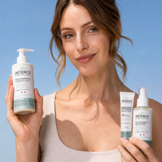 Femme tenant les trois soins des Essentiels de l'Été Réjence devant un ciel bleu : Soin Lavant Apaisant, Gel-Crème Hydratant et Soin Correcteur de Teint SPF30 pour une routine estivale complète des peaux sensibles.
