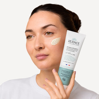 Femme tenant la Cold cream SOS rougeurs et démangeaisons 150 mL Réjence avec une noisette de crème appliquée sur la joue.