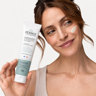 Femme tenant la Crème riche SOS sécheresses et squames 150 mL Réjence avec une noisette de crème appliquée sur la joue.