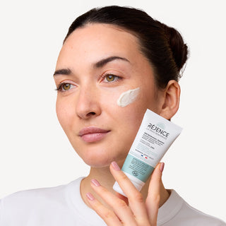 Femme tenant la Crème riche SOS sécheresses et squames Réjence avec une noisette de crème appliquée sur la joue.
