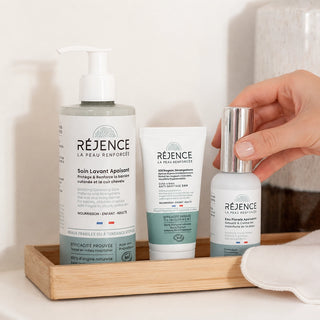 Les soins de la routine anti-rougeurs Réjence disposés sur un plateau dans une salle de bain, avec une main saisissant l'eau florale apaisante.