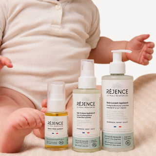 Les trois soins Réjence de la routine prévention eczéma bébé posés devant un bébé assis sur une serviette beige, illustrant une routine de soin quotidienne pour peau atopique.